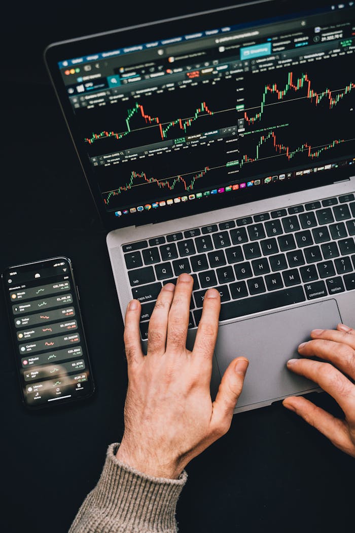 hero-img-01 Hands typing on a laptop with cryptocurrency charts and smartphone display in view, focusing on digital finance.