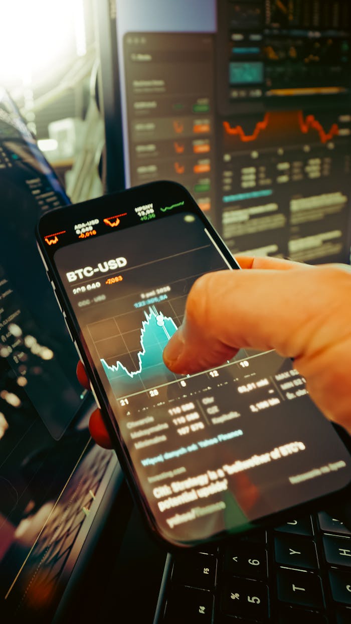 hero-img-02 A hand holds a smartphone displaying BTC-USD chart amidst digital trading screens, highlighting crypto finance.